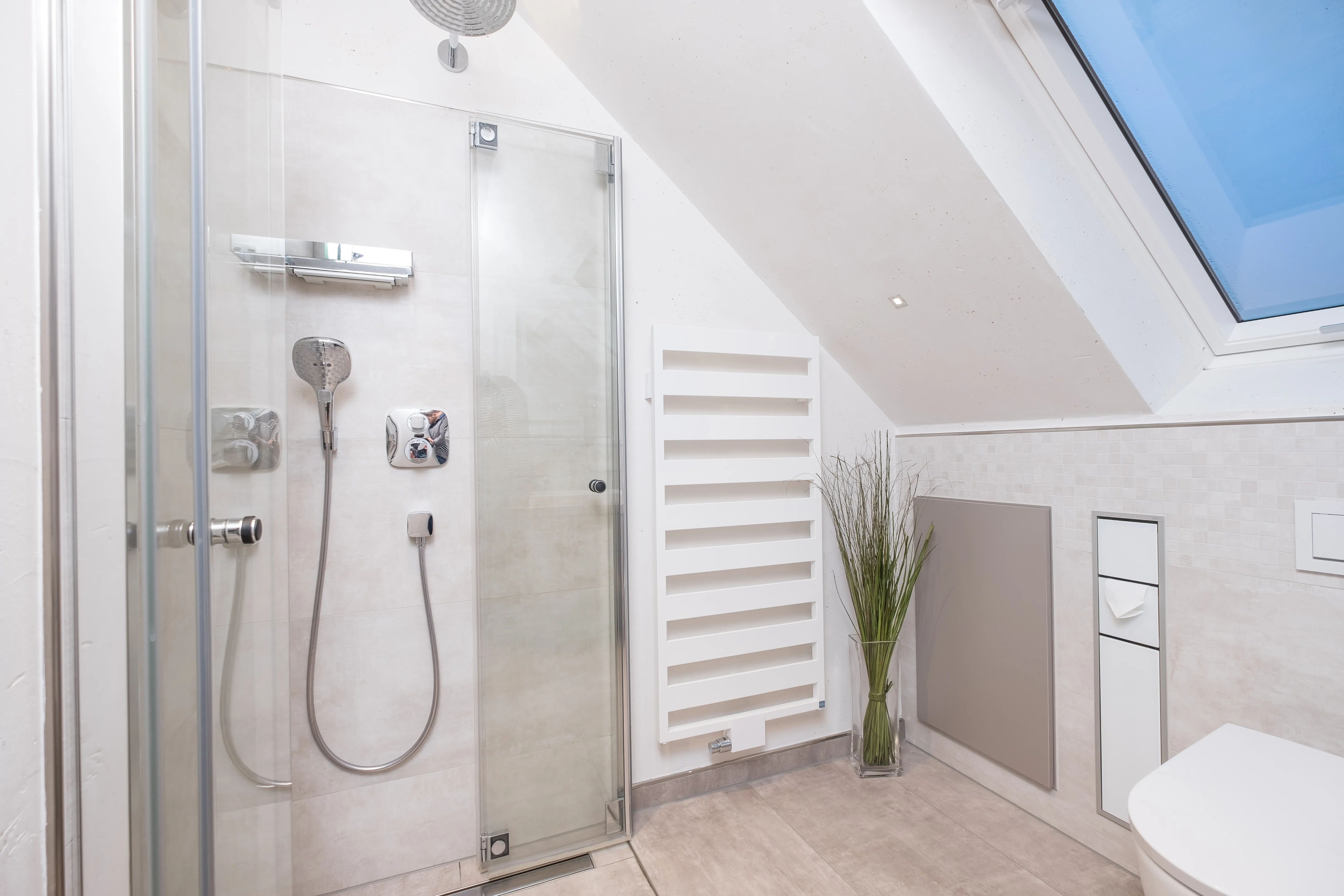 Bodengleiche Eckdusche unter Dachschräge mit Glasabtrennung, Regendusche und weißem Design-Heizkörper.