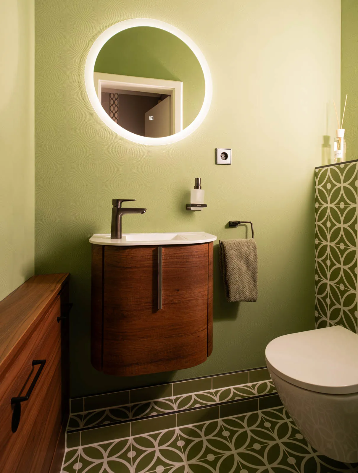 Waschbecken im Gäste-WC mit rundem LED-Spiegel und grüner Wandgestaltung.