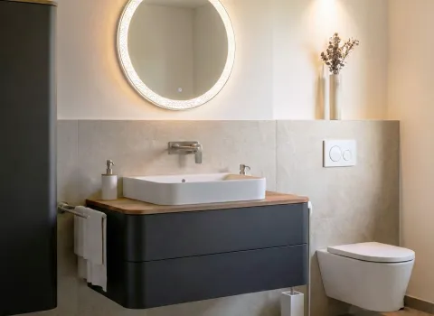 Fotorealistischer Planungsentwurf eines kompakten Comfort-Badezimmers mit Waschtisch, rundem Leuchtspiegel und schwebender Toilette in gedimmten Licht.