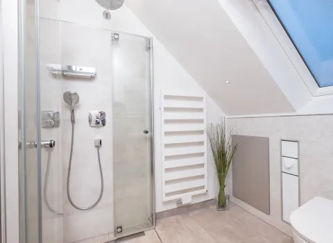 Bodengleiche Eckdusche unter Dachschräge mit Glasabtrennung, Regendusche und weißem Design-Heizkörper.