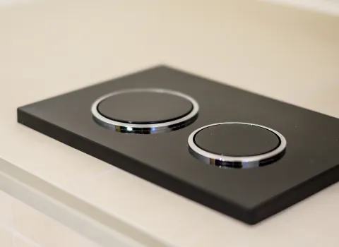 Detailaufnahme einer schwarzen WC-Drückplatte mit zwei runden Tasten in einem modernem Badezimmer.