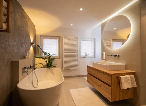 Modernes Badezimmer mit freistehender Badewanne, Holzwaschtisch und rundem LED-Spiegel.