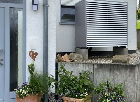 Außengerät einer Wärmepumpe an der Hausfassade mit Gartenbepflanzung.